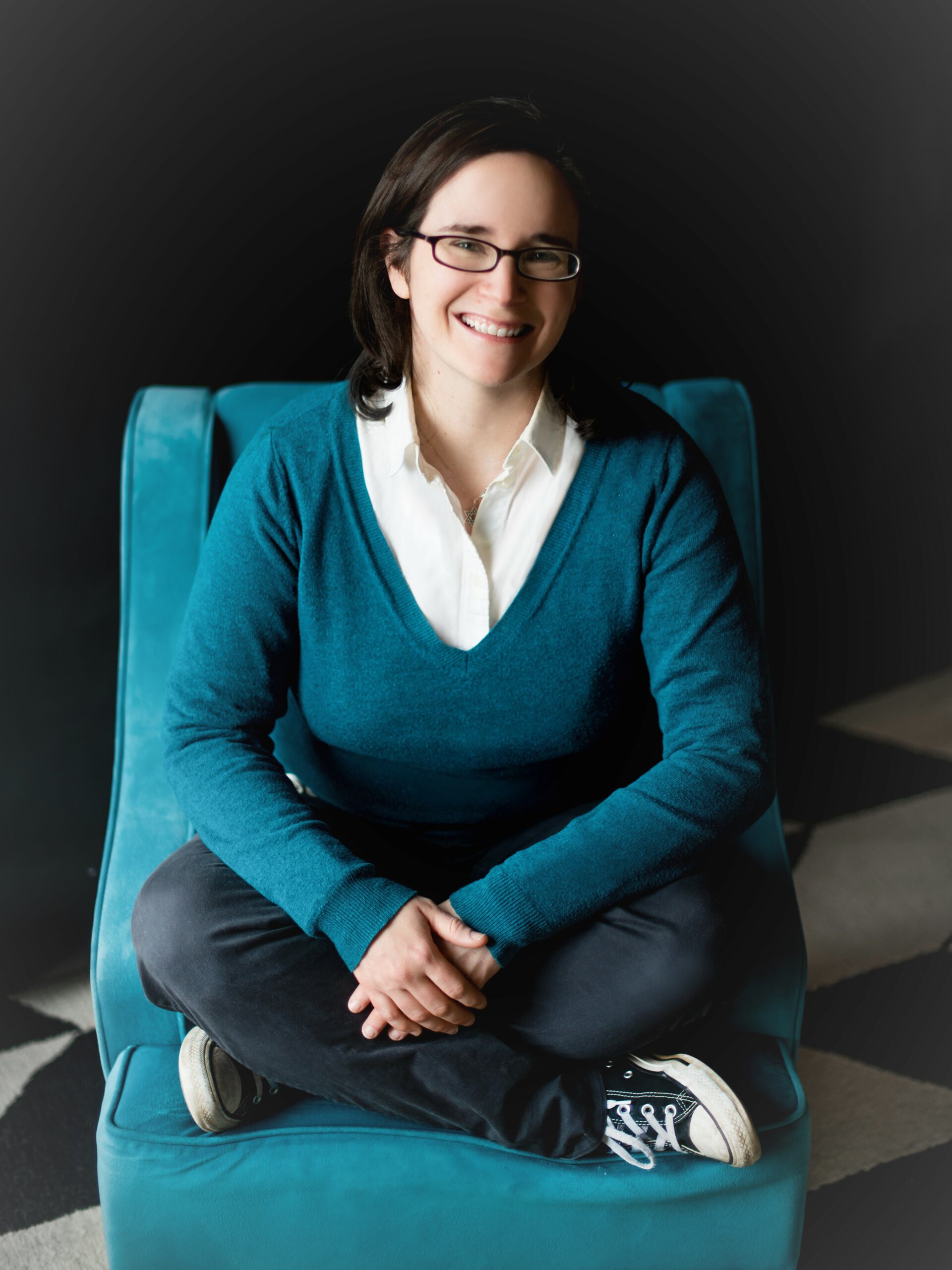 A smiling woman dressed in blue sitting cross-legged on a blue chair